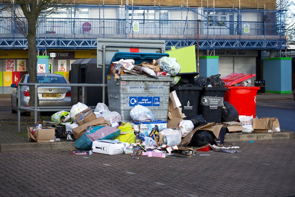 A large collection of mixed household waste, including crumpled cardboard boxes, black and red rubbish bags, and scattered paper and packaging, is piled around a grey recycling bin with a blue lid on 