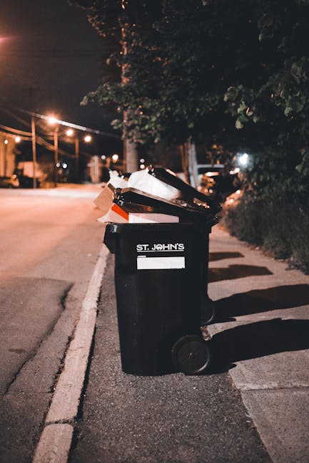 A large collection of mixed household waste, including crumpled cardboard boxes, black and red rubbish bags, and scattered paper and packaging, is piled around a grey recycling bin with a blue lid on 