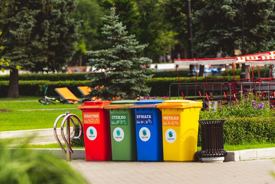 An outdoor scene showing a pile of mixed waste materials arranged on a paved surface, possibly for collection or disposal. The heap includes various items such as discarded cardboard boxes, plastic co